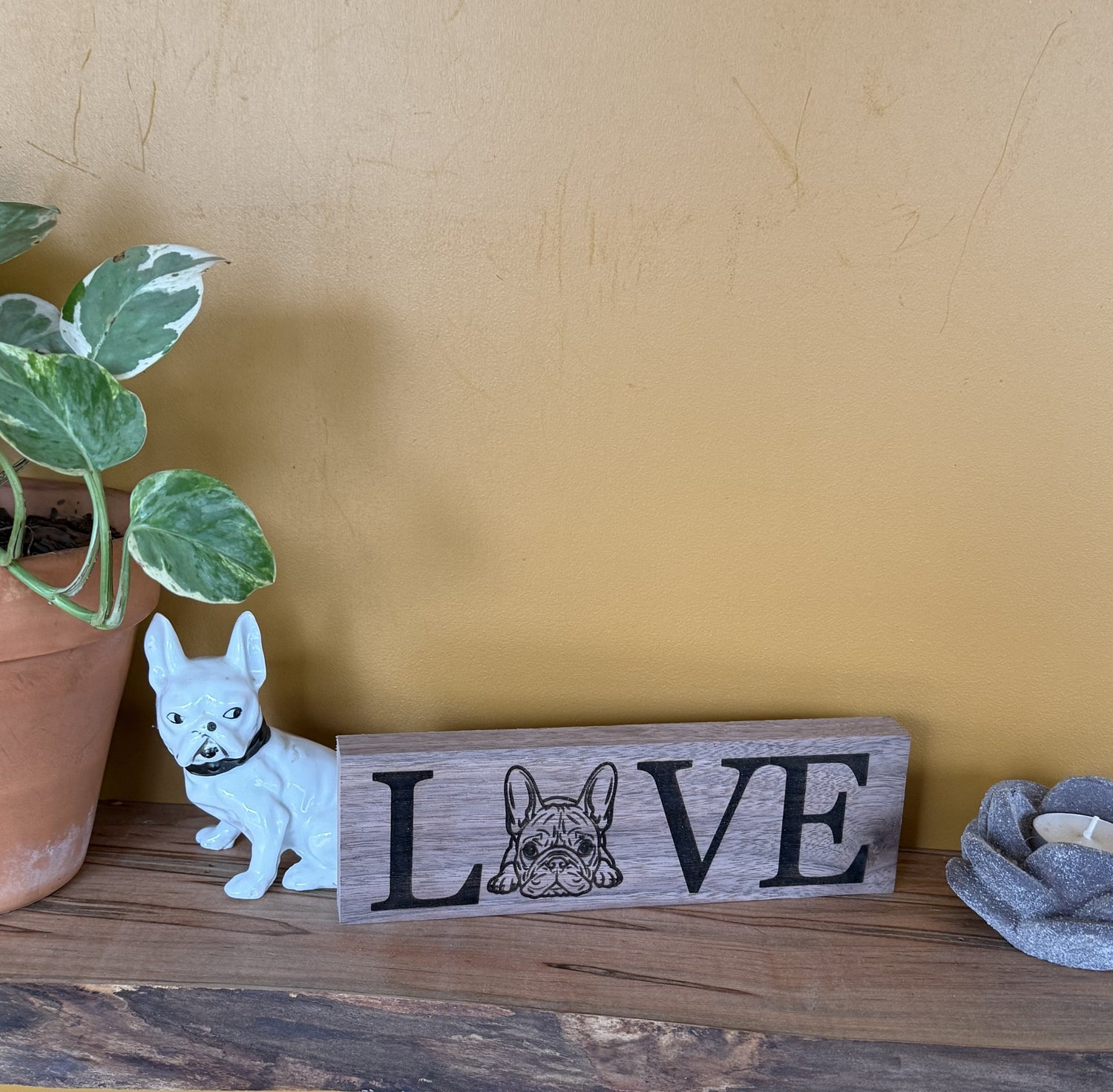 Wooden sign with 'LOVE' and a French Bulldog illustration on a wooden surface with a yellow wall background.