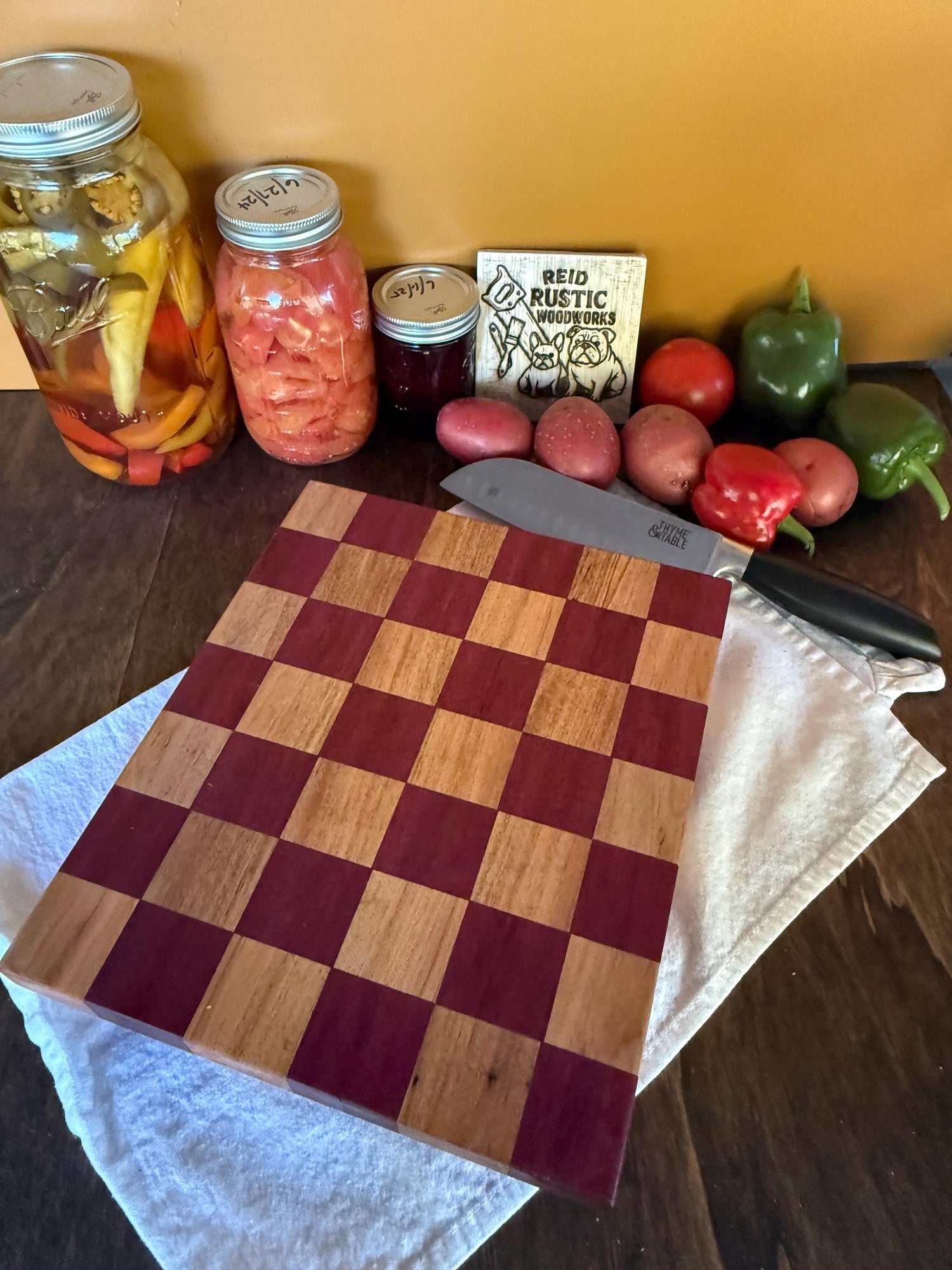 Handmade checkered cutting board made with purpleheart and maple hardwood on a wooden table with jars, vegetables, and a knife.