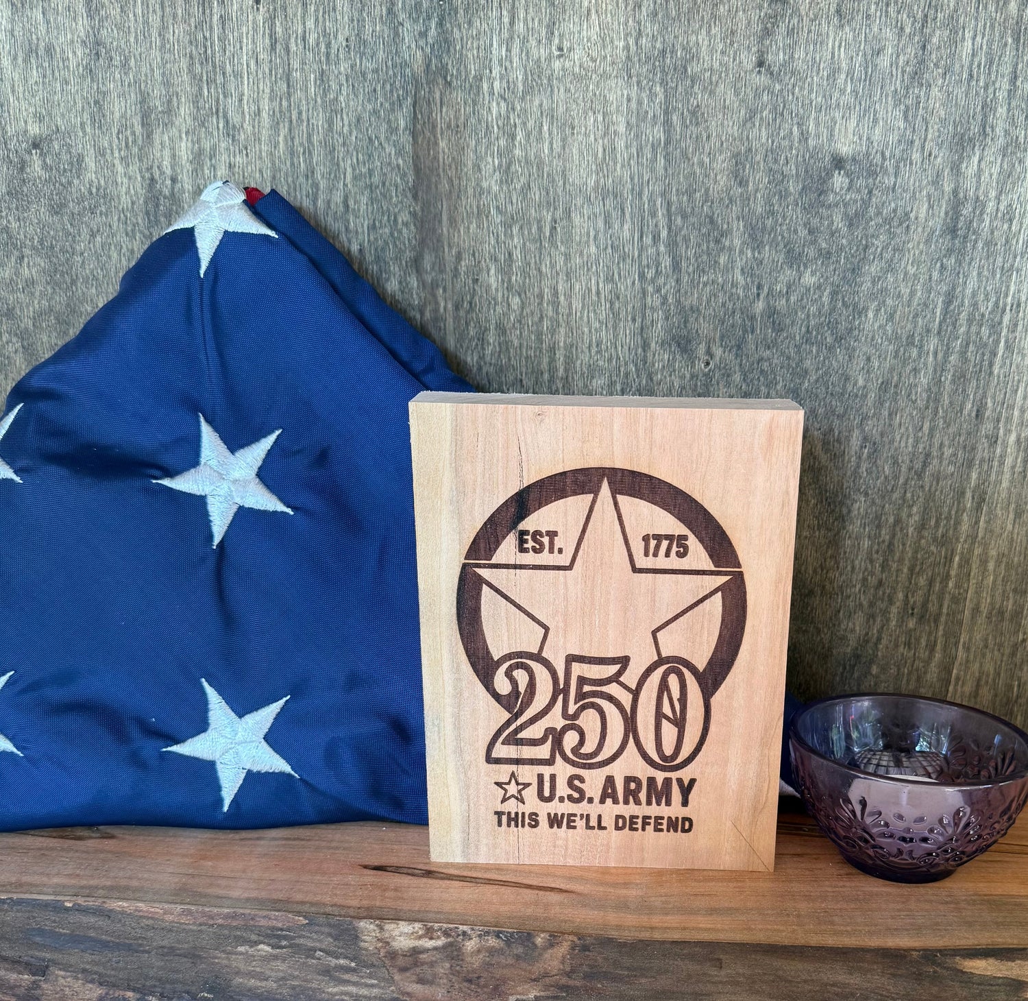 Wooden plaque with U.S. Army 250th anniversary design next to a flag on a wooden surface.