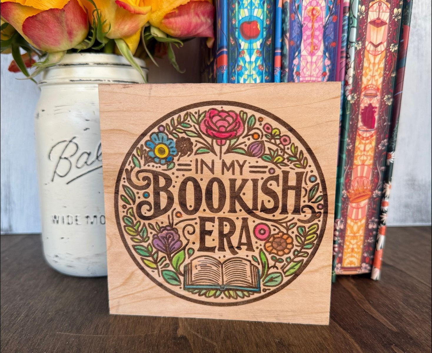 Wooden handpainted block with 'In My Bookish Era' design, next to a vase of flowers