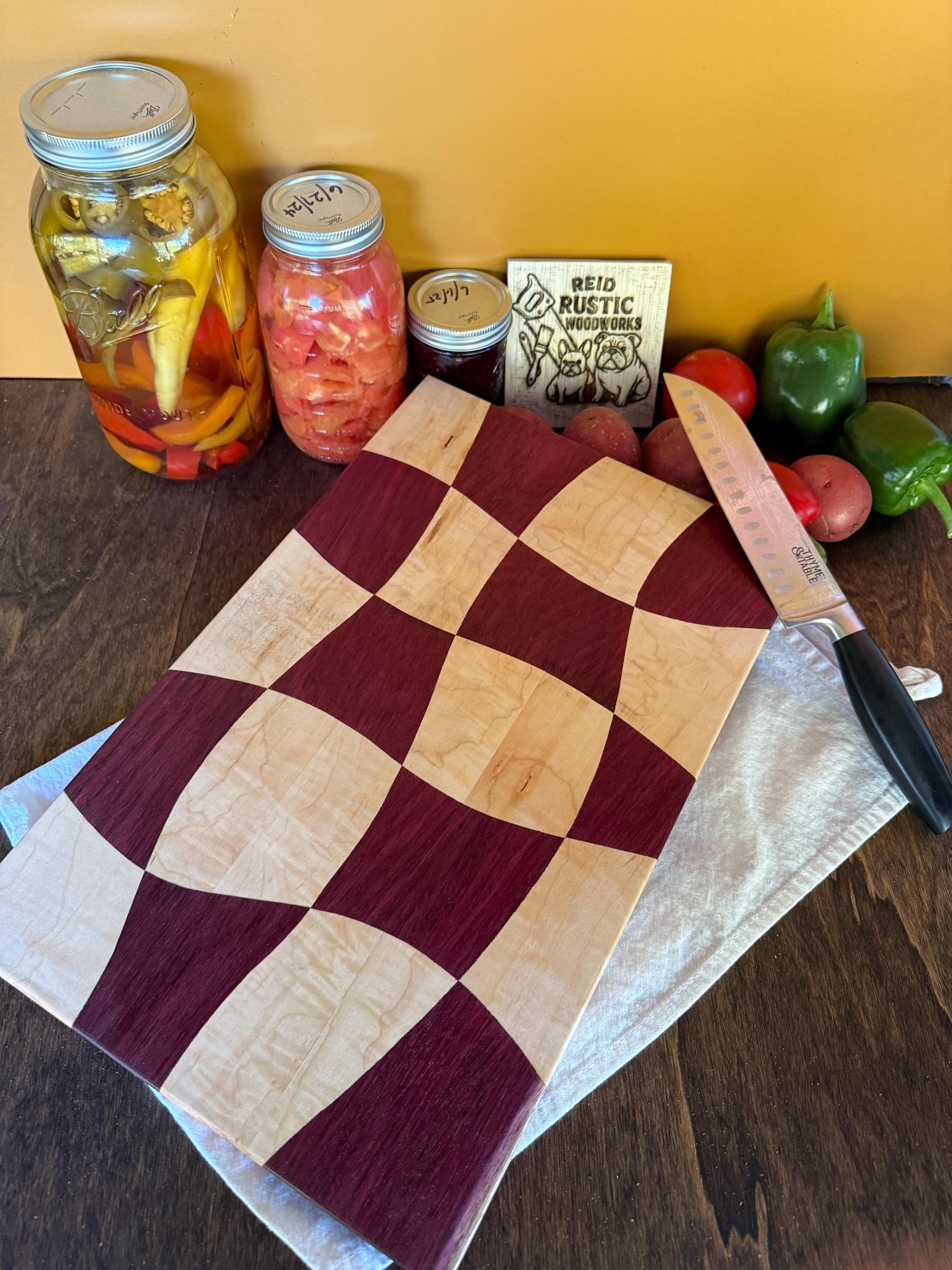 Wavy Checkered cutting board on a wooden surface with jars, vegetables, and a knife.