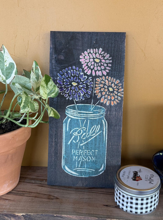 Decorative sign with a mason jar design on a wooden surface next to a plant and small container.