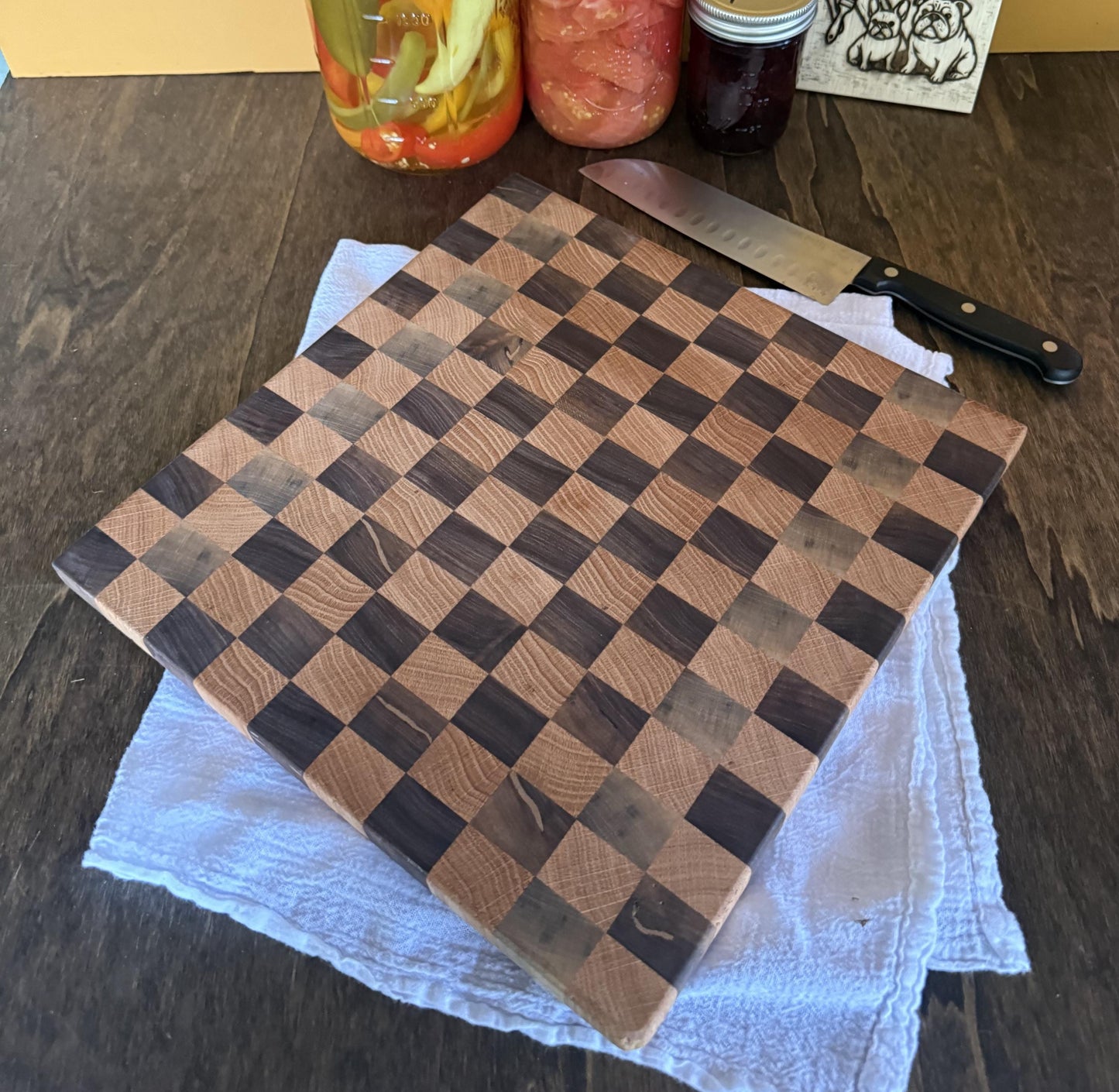 Checkered wooden cutting board on a dark surface with a knife and jars in the background.
