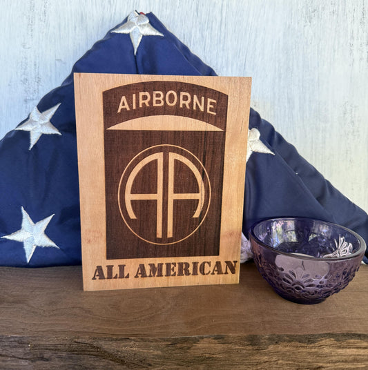 Wooden plaque with 'Airborne All American' engraving in front of a folded American flag on a wooden surface.