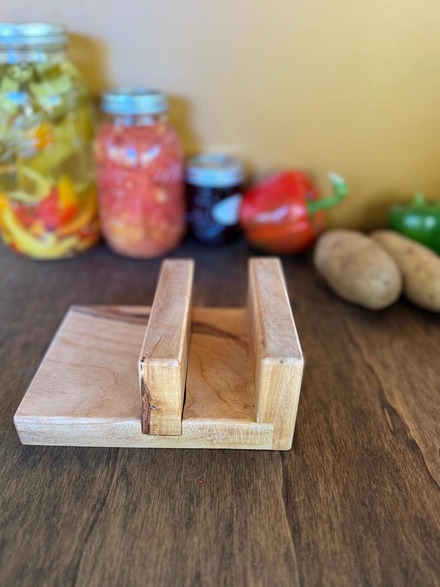 Wooden cutting board holder on a wooden surface with jars and vegetables in the background.