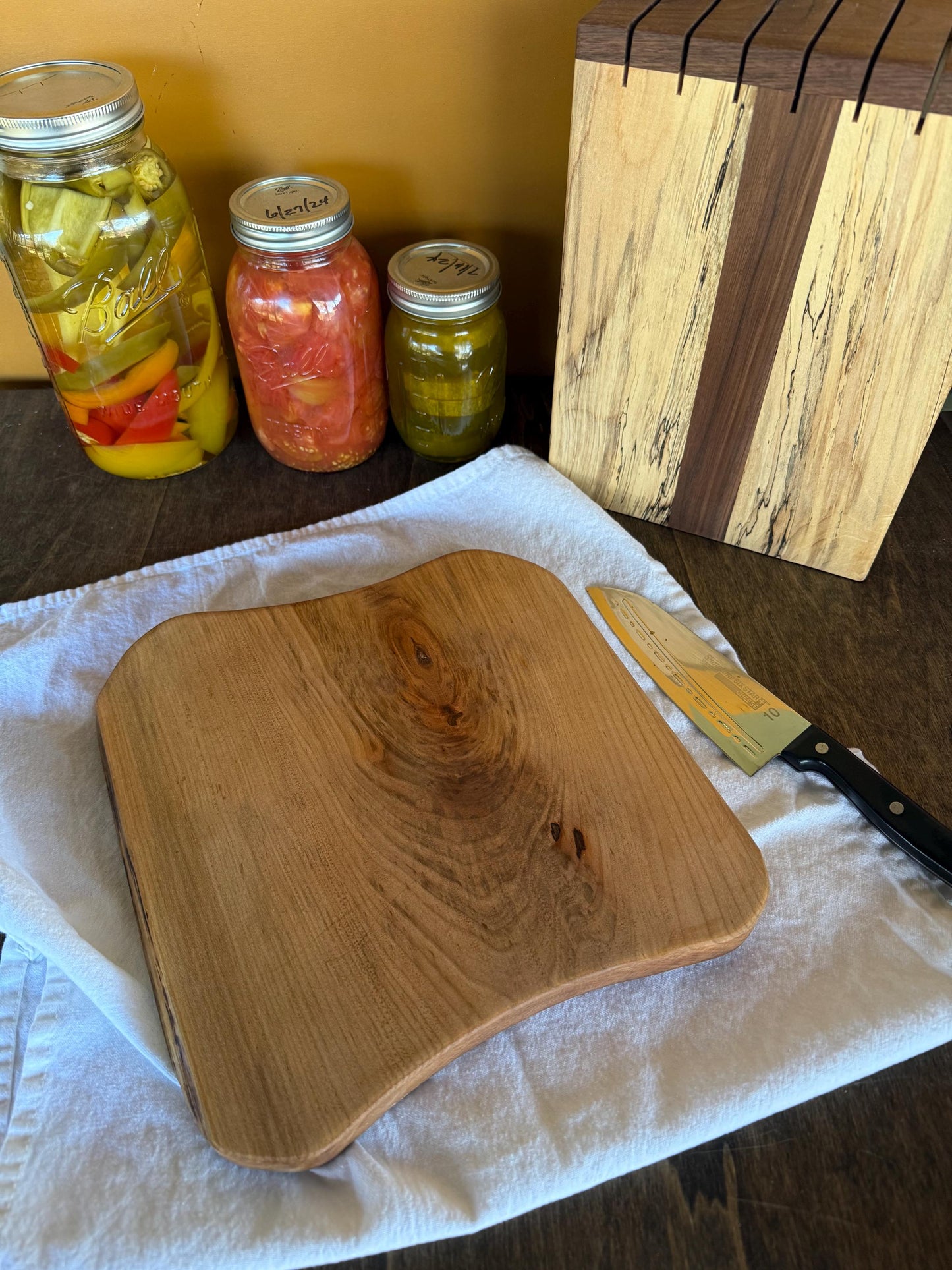 Wooden cutting board on a towel with jars of pickled vegetables in the background.