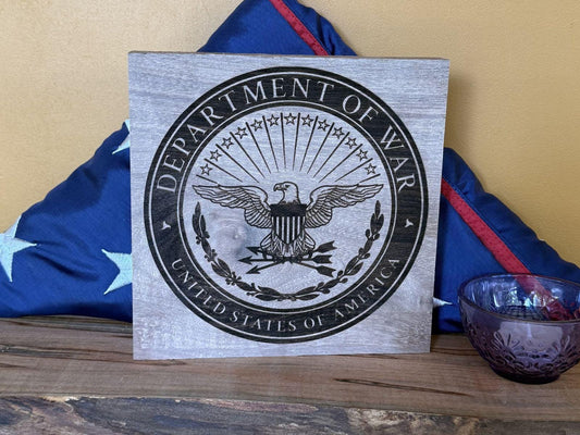 Wooden plaque with Department of War emblem on a wooden surface with a flag in the background