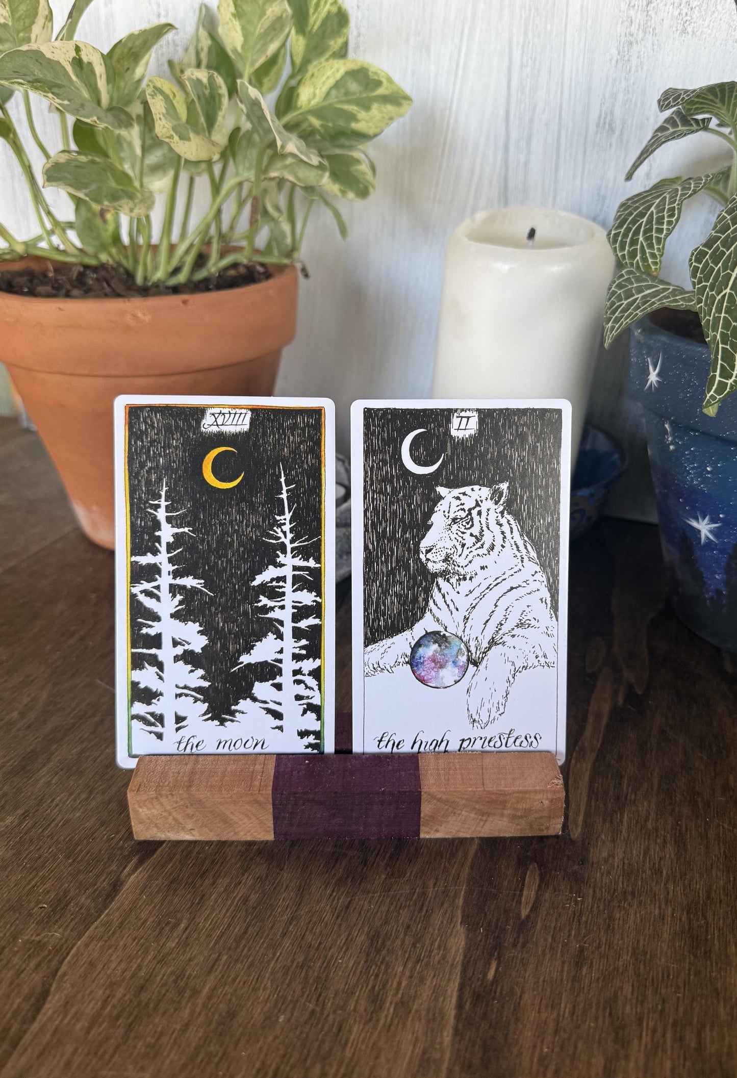 Two tarot cards on a wooden stand with plants and a candle in the background.