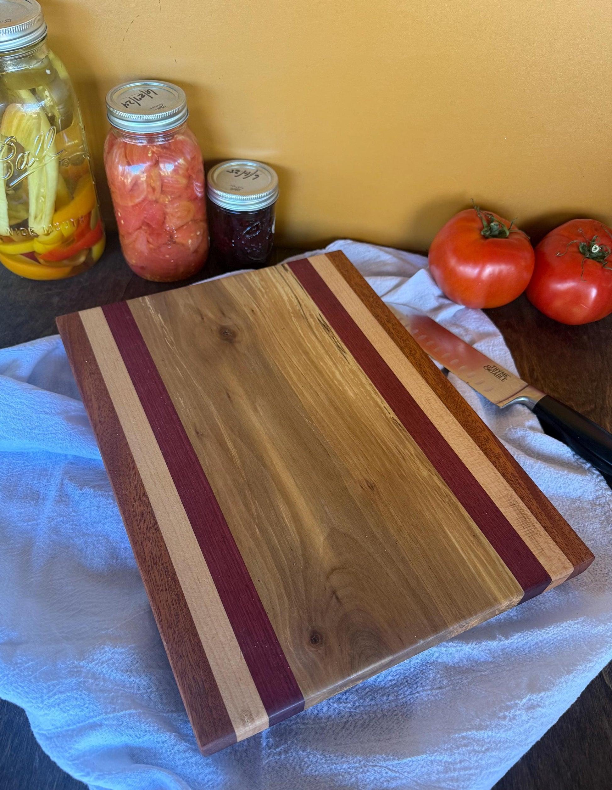 Wooden cutting board with a knife on a table with tomatoes and jars in the background.