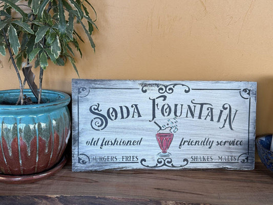Wooden sign with 'Soda Fountain' text and graphics on a wooden surface with a plant in the background.
