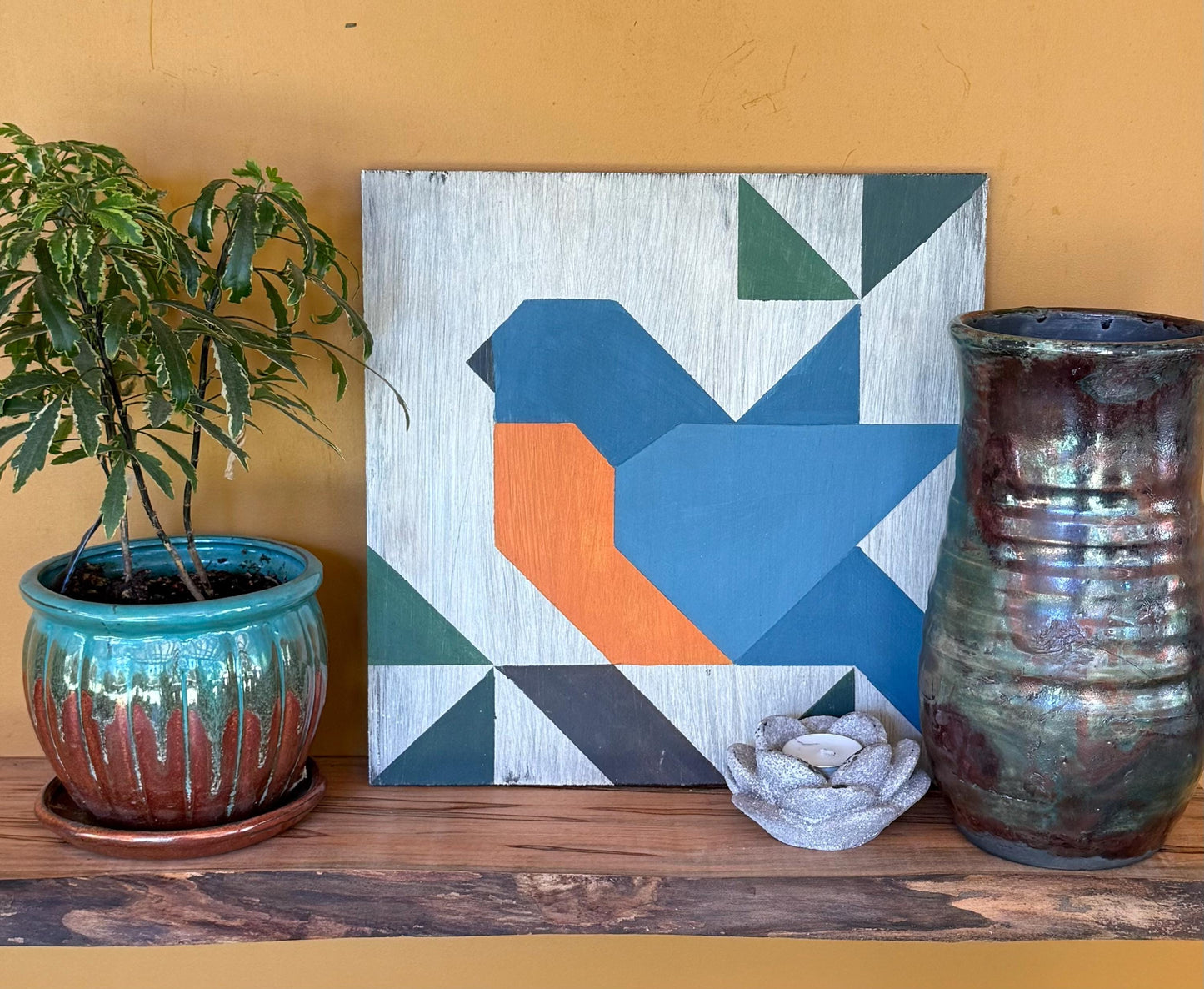 Geometric barn quilt with an abstract Blue Bird motif on a wooden shelf with plants, and a vase against a yellow wall.