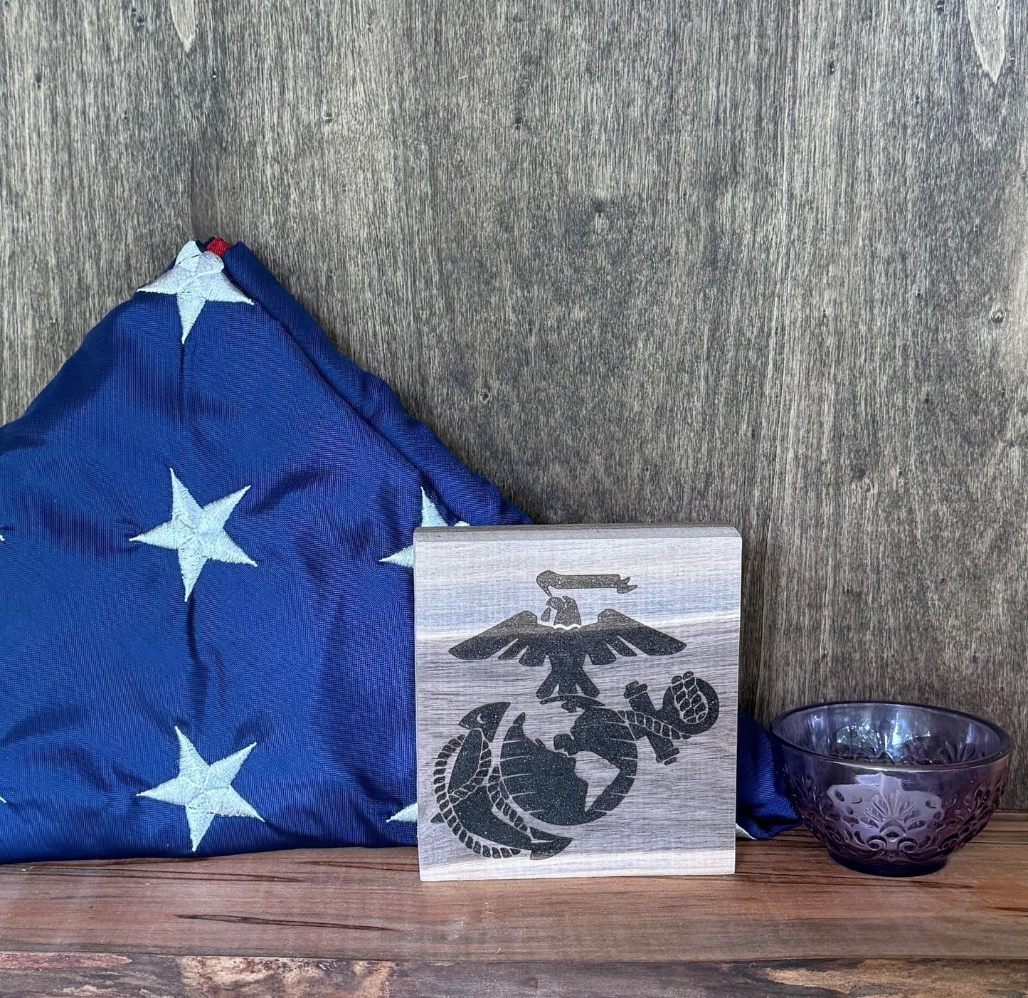 Wooden plaque with USMC logo next to a blue flag and purple bowl on a wooden surface.
