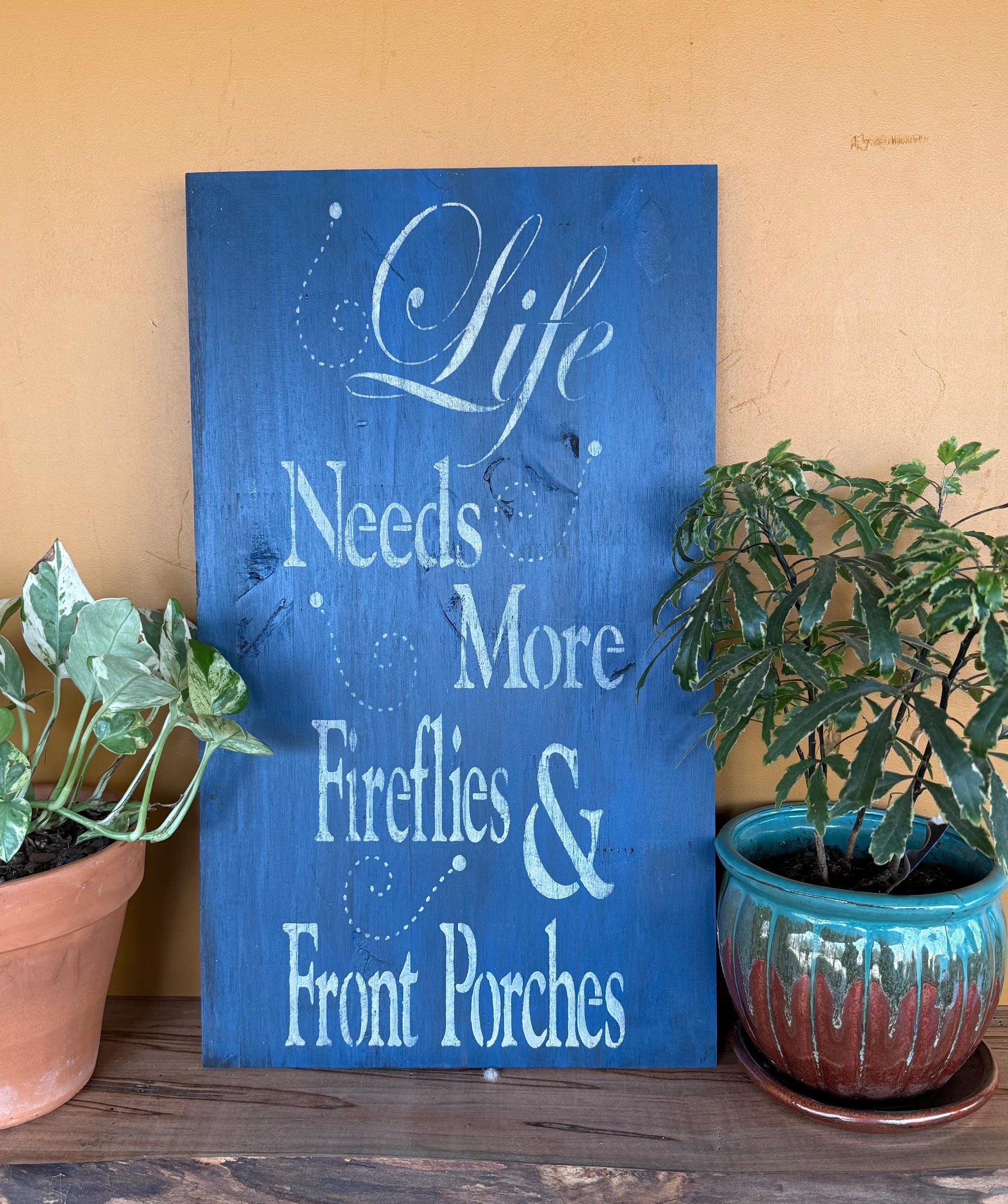 Blue sign with white text 'Life Needs More Fireflies & Front Porches' on a wooden surface with potted plants.