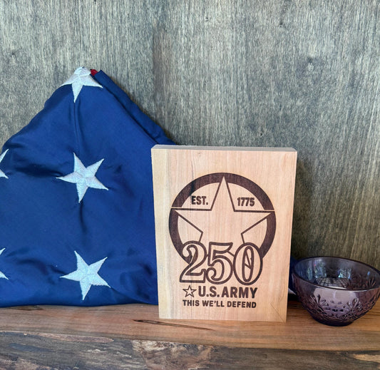 Wooden plaque with U.S. Army 250th anniversary design next to a flag on a wooden surface.