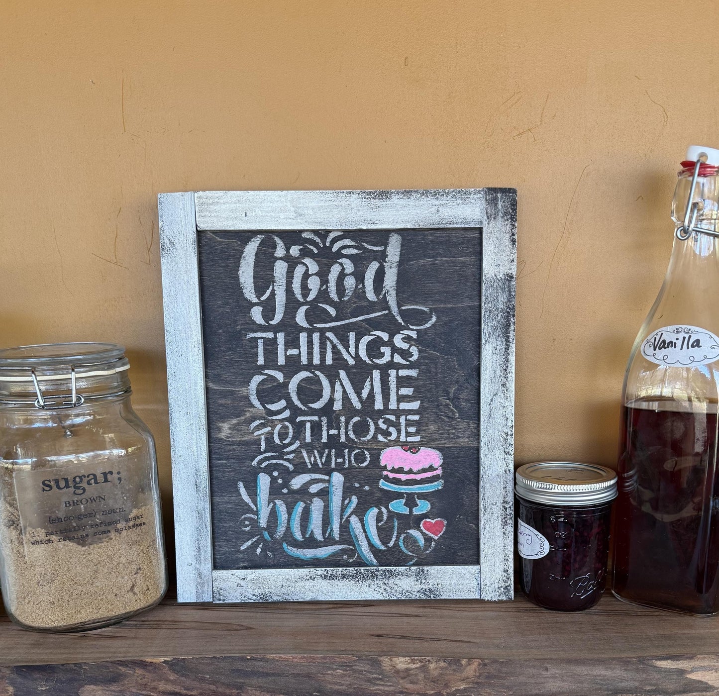 Decorative sign with 'Good things come to those who bake' text, surrounded by jars and a bottle on a wooden surface.