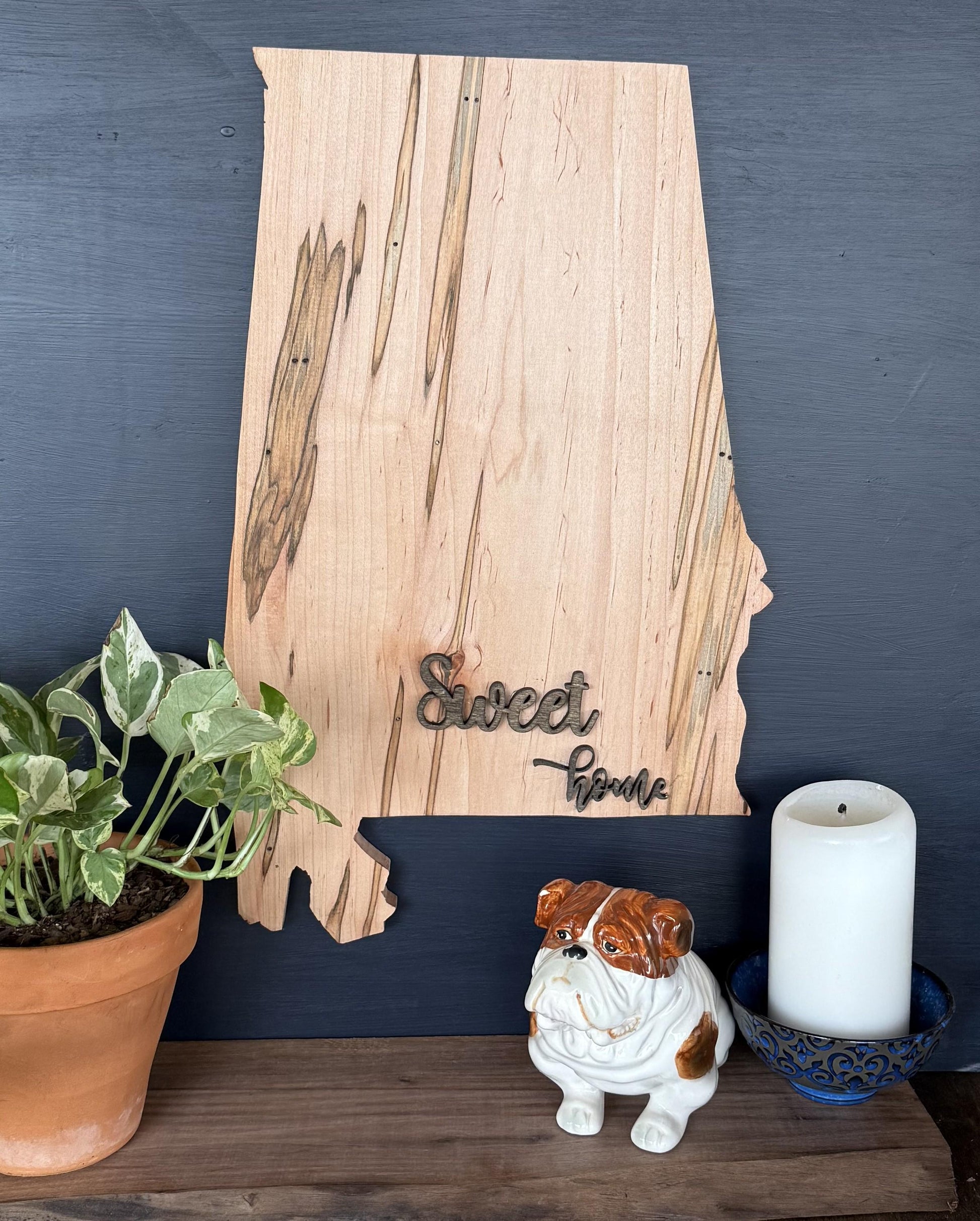 Wooden Alabama shaped sign with 'Sweet Home' text, potted plant, ceramic bulldog figurine, and candle on a dark surface.