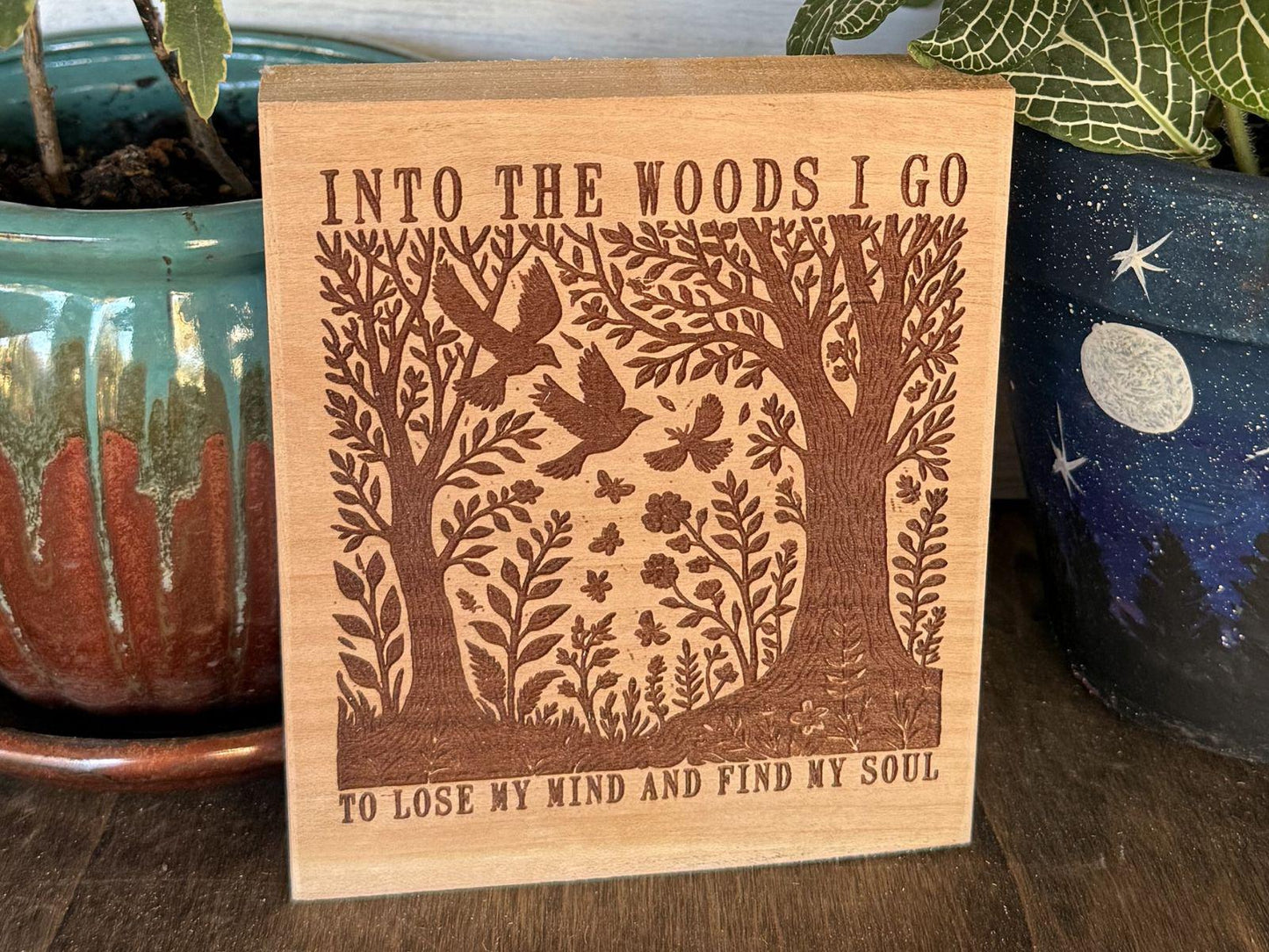 Wooden sign with nature-themed design and text that says "Into the Woods I Go, to Lose my Mind and Find my Soul", placed between two potted plants.
