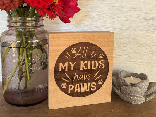 Wooden sign with 'All My Kids Have Paws' text next to a vase of red flowers and a candle.