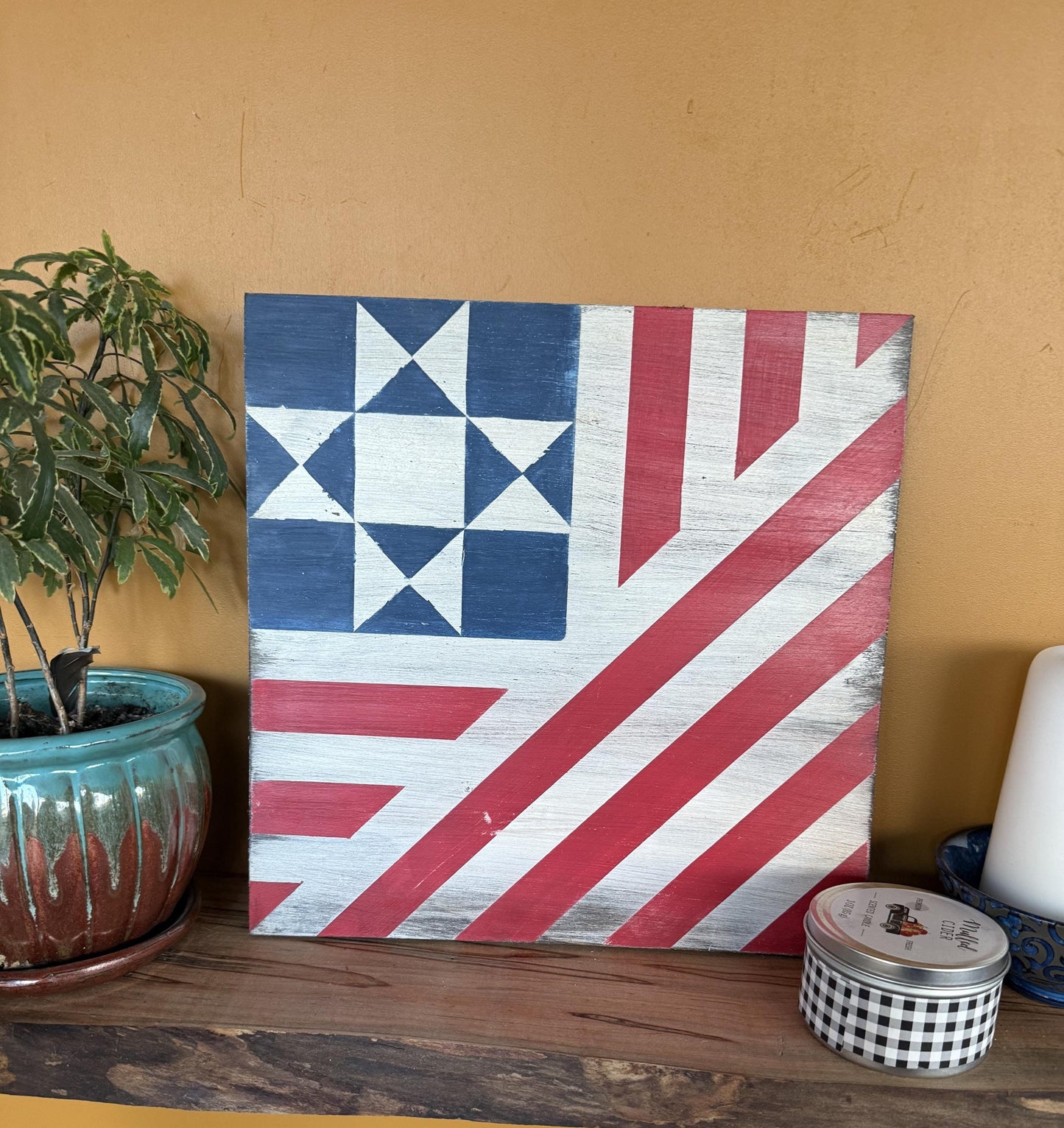 Barn Quilt with a geometric design resembling a flag on a shelf with a plant and candle.