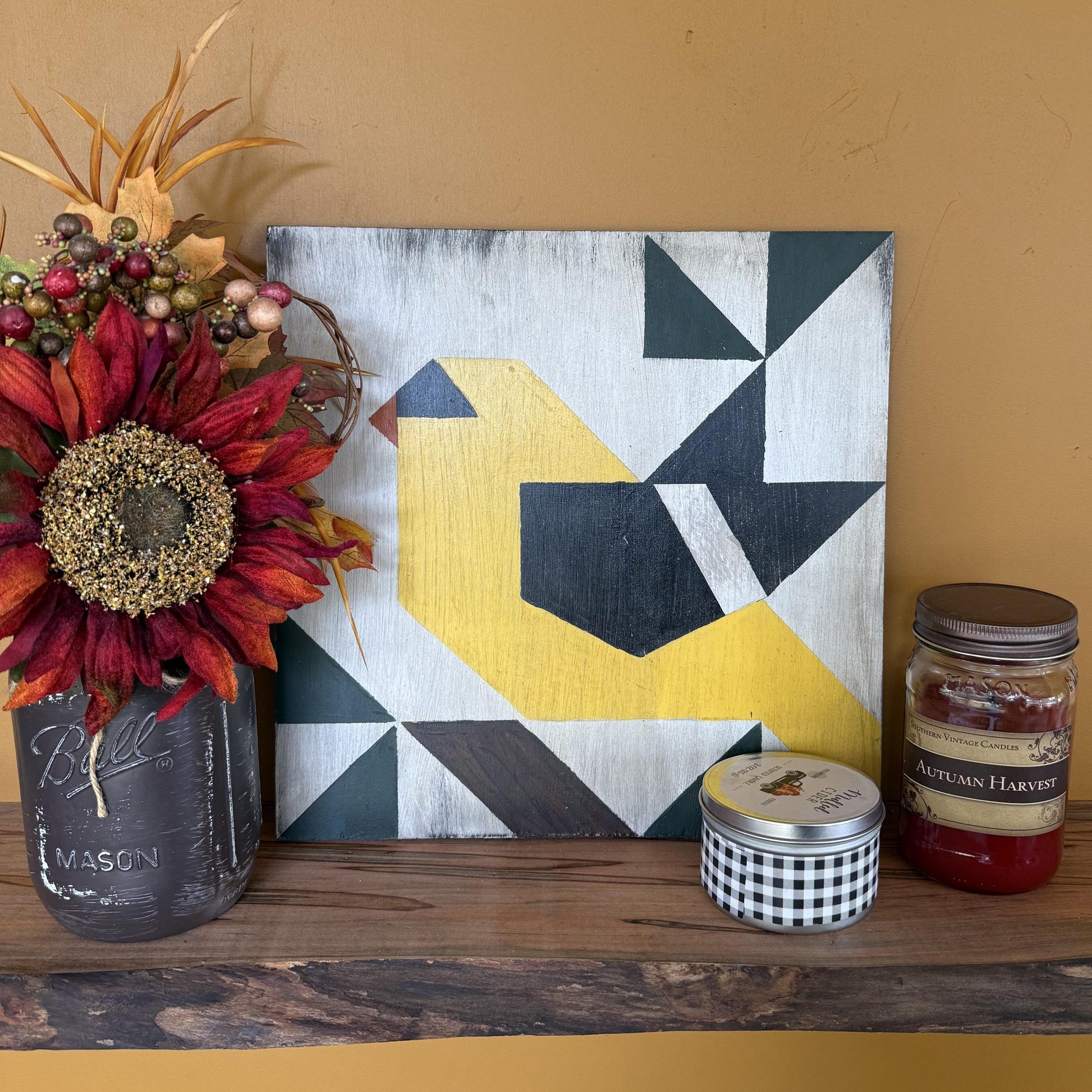 Geometric barn quilt with an abstract Goldfinch motif on a wooden shelf with a plant and candles.