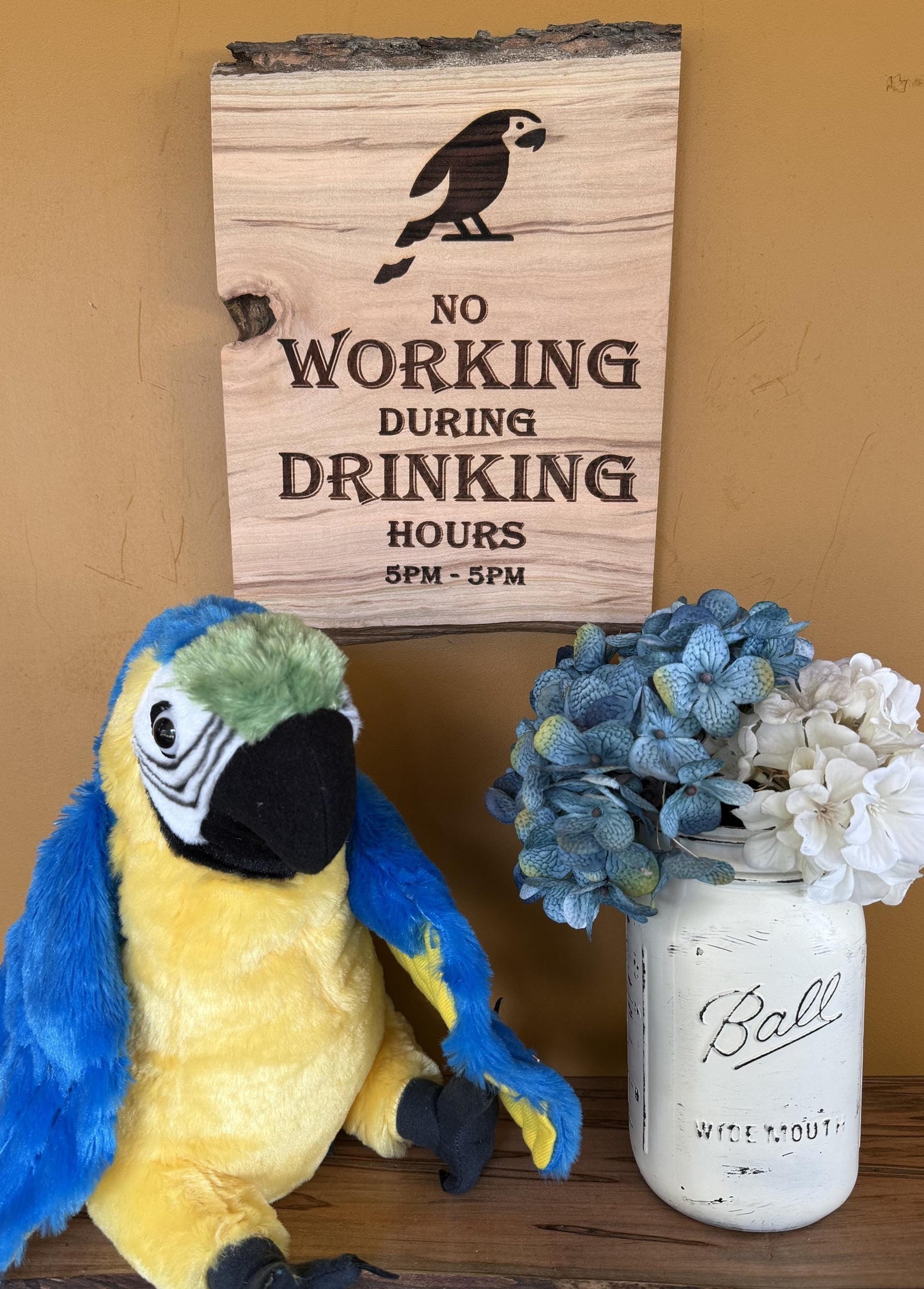 Wooden sign with parrot illustration and text that says No Working During Drinking Hours 5pm to 5pm, plush parrot toy, and floral arrangement on a wooden surface.