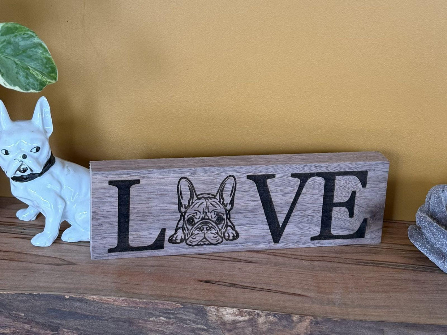 Wooden sign with 'LOVE' and a French Bulldog illustration on a wooden surface with a yellow wall background.