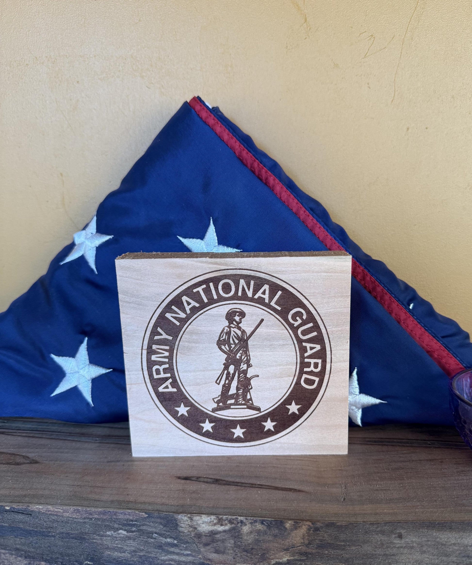 Wooden sign with Army National Guard emblem in front of a folded American flag on a wooden surface.