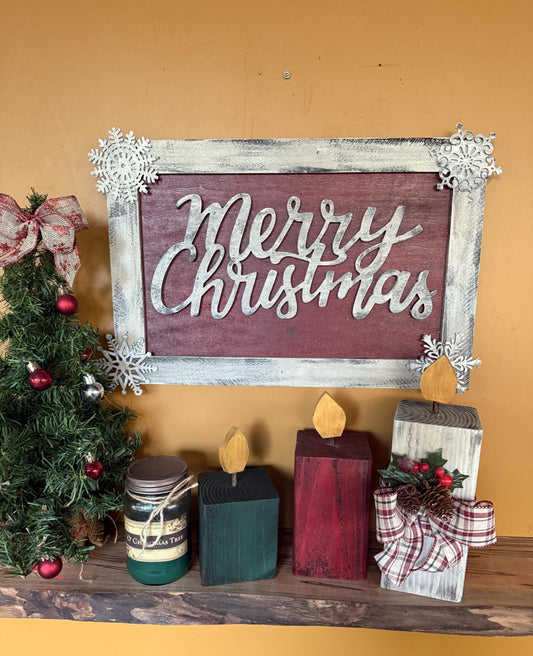 Decorative Christmas sign with 'Merry Christmas' on a wooden frame, surrounded by candles and a small tree.