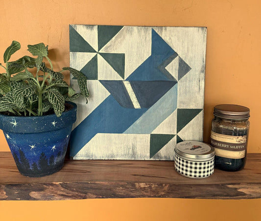 Geometric barn quilt with an abstract Blue Jay motif on a wooden shelf with a plant and candles.