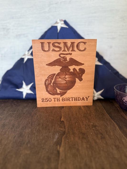 Wooden plaque with USMC logo and text on a wooden surface with an American flag in the background.