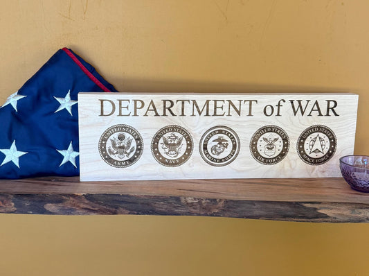 Wooden sign with 'DEPARTMENT of WAR' and military emblems on a shelf with an American flag in the background.