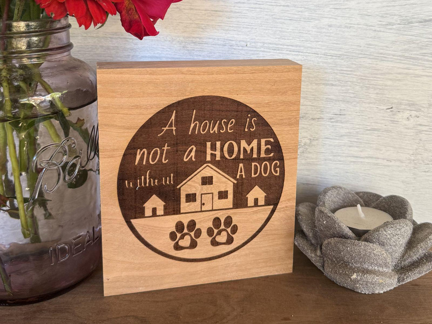 Wooden sign with 'A house is not a home without a dog' text, next to a mason jar with flowers and a candle.