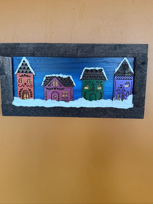 Painting of four colorful houses on a wooden panel against a beige wall.