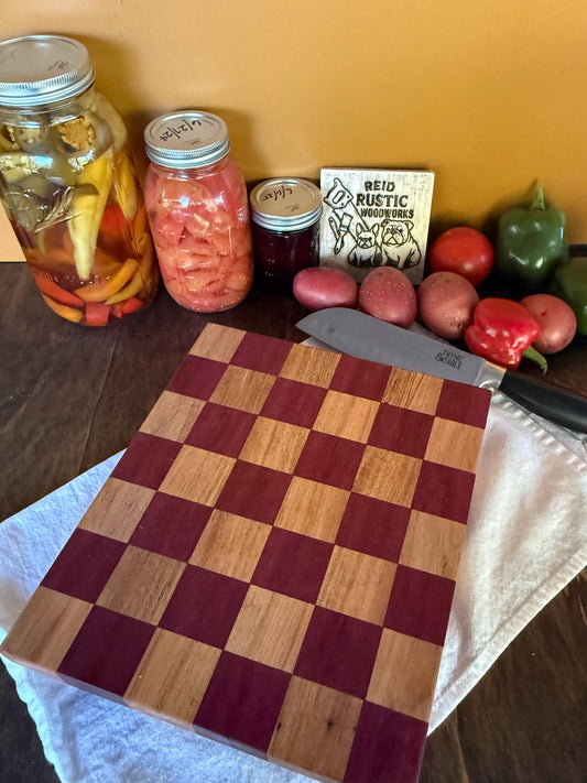 Handmade checkered cutting board made with purpleheart and maple hardwood on a wooden table with jars, vegetables, and a knife.