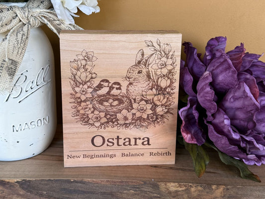 Wooden block with Ostara design featuring a rabbit, birds, and flowers, placed next to a mason jar and purple flowers.