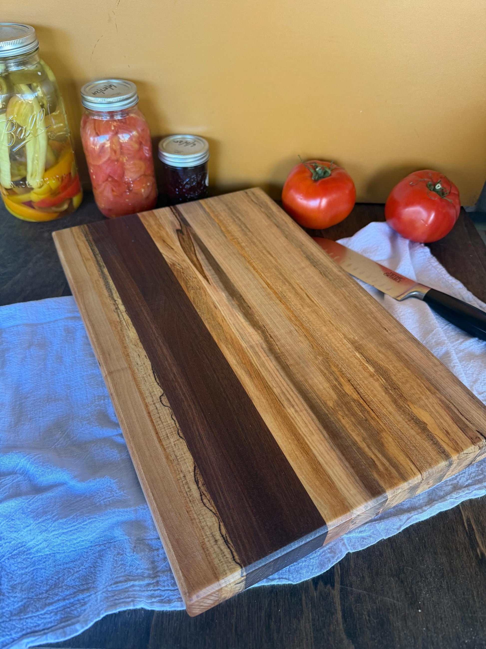 Wooden cutting board on a table with tomatoes and jars in the background