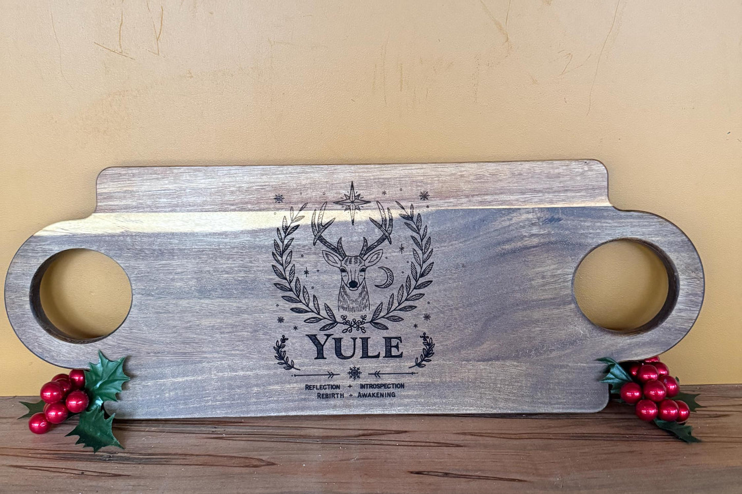 Wooden cutting board with deer design and 'Yule' text, surrounded by holly and berries on a wooden surface.
