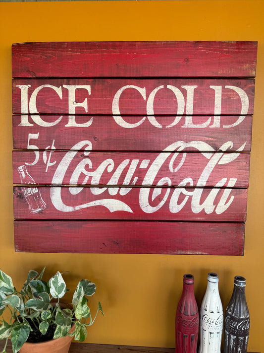 Wooden sign with 'Ice Cold Coca-Cola' on a yellow wall with decorative bottles below.