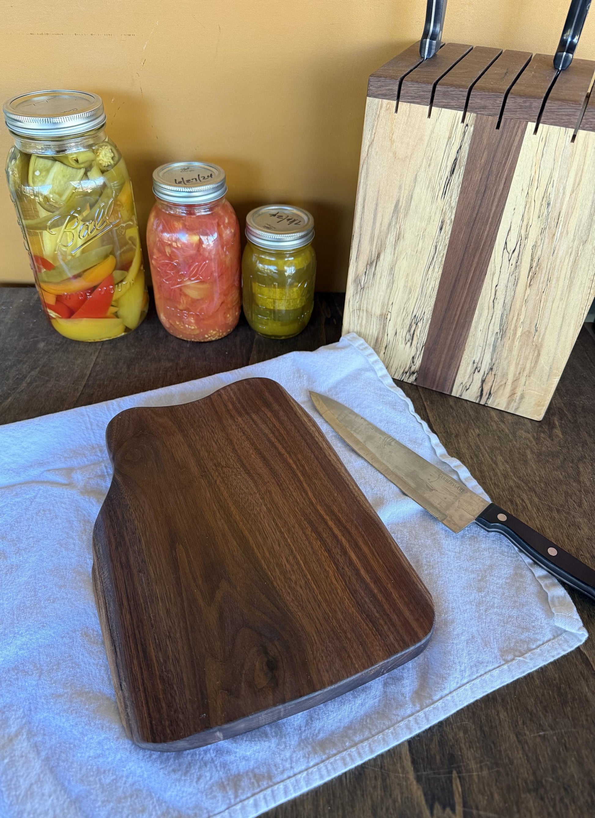 Wooden cutting board with a knife on a towel, jars of pickled vegetables in the background.