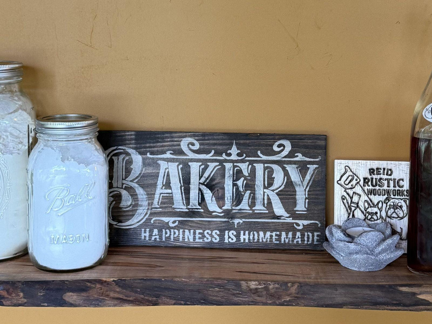 Wooden shelf with 'Bakery' sign, jars, and decorative items against a beige wall.