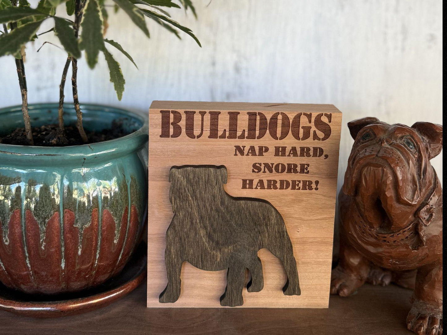 Wooden sign that says "Bulldogs  Nap Hard, Snore Harder" next to a potted plant and another bulldog figurine.