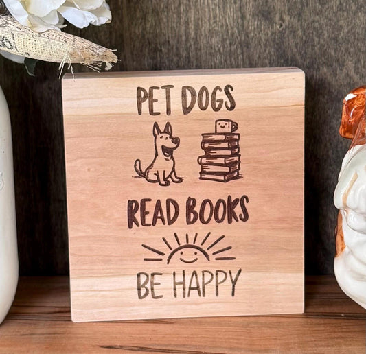 Wooden sign with text 'Pet dogs, read books, be happy' on a wooden shelf.