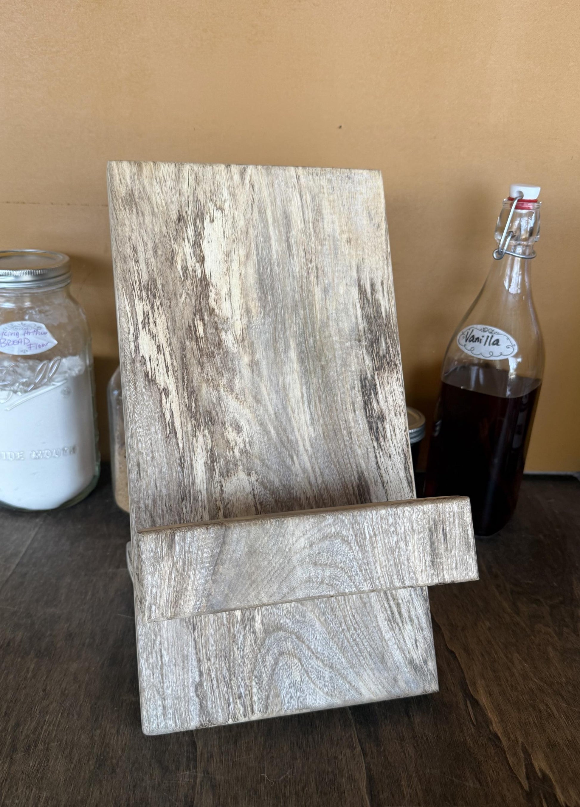 Wooden cookbook stand on a wooden surface with jars and a bottle in the background