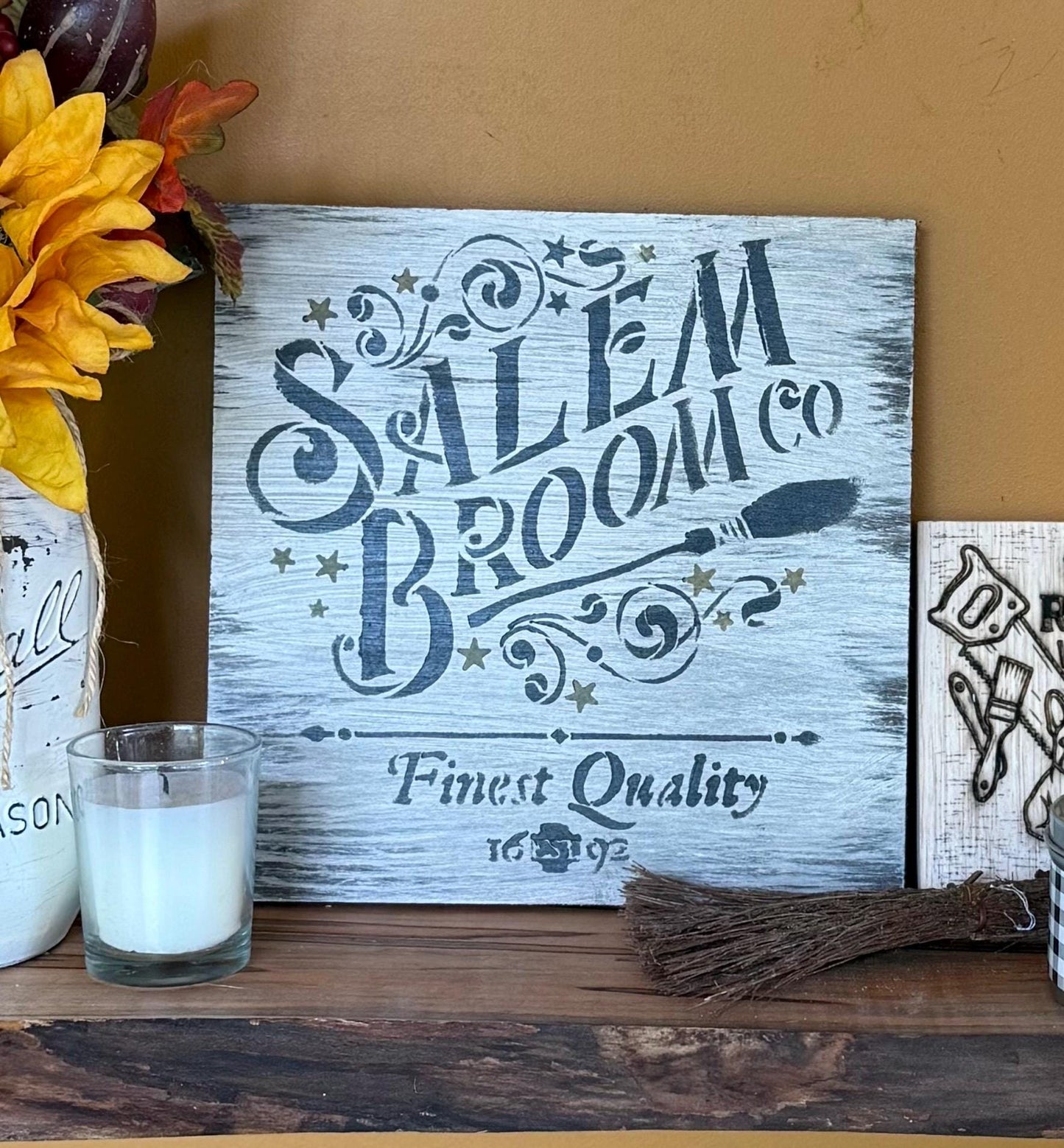 Decorative sign with 'Salem Broom Co. Finest Quality' text on a wooden surface with flowers and a candle.