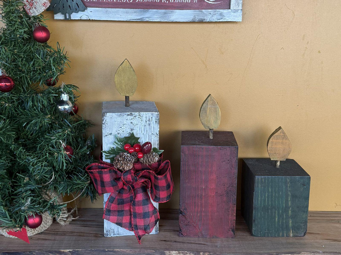 Wooden candles with a plaid bow next to a Christmas tree against a yellow wall.