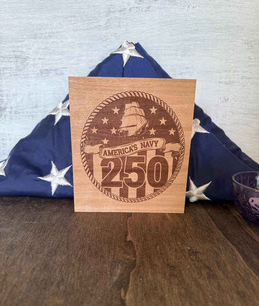 Wooden plaque celebrating America's Navy 250th anniversary with an American flag in the background.