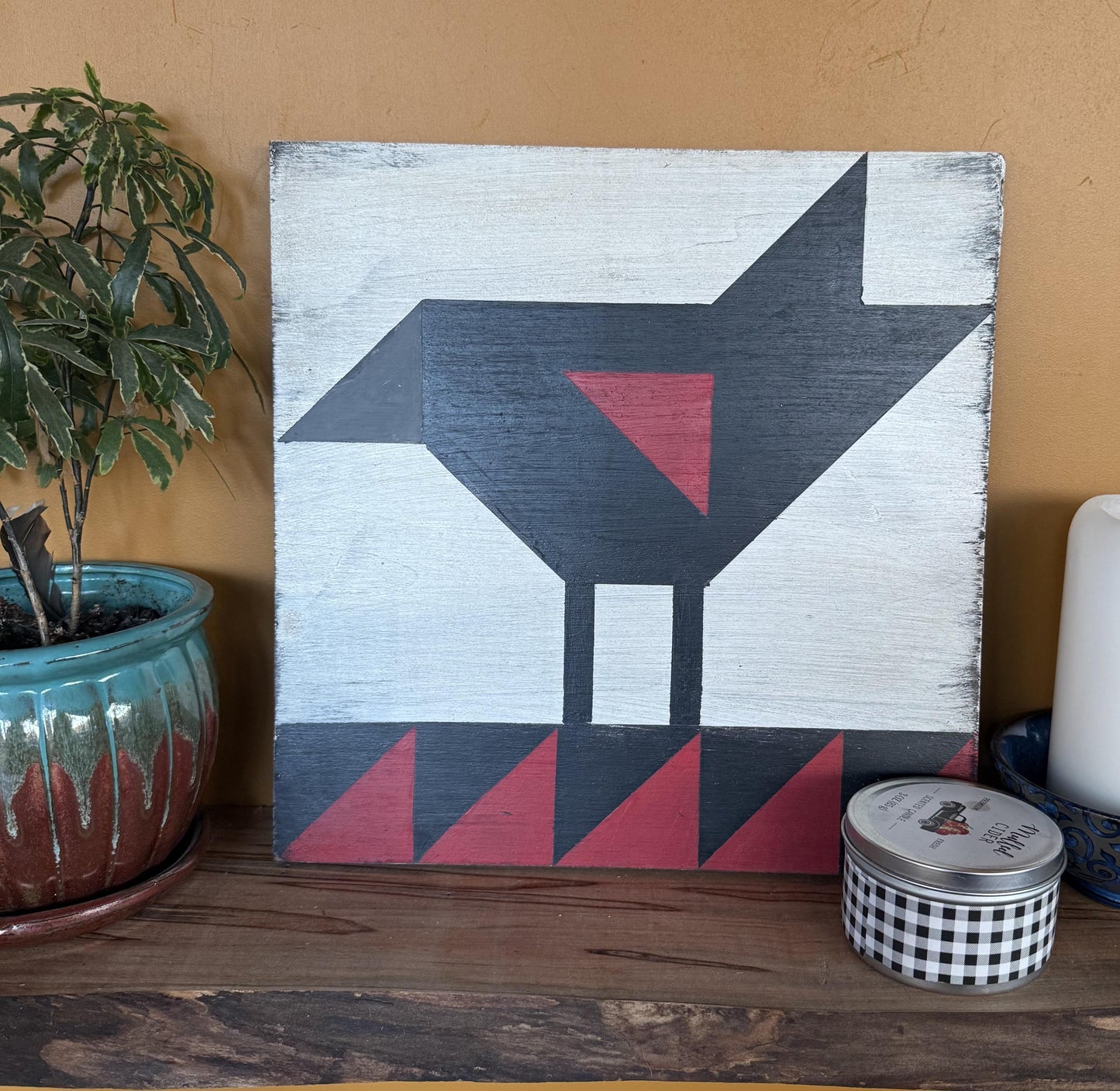 Geometric barn quilt with an abstract Red wing black bird motif on a wooden shelf with a plant and candles.