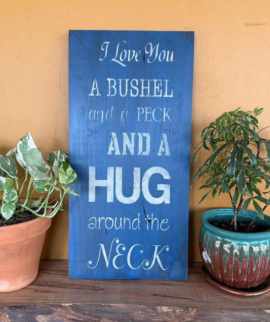 Decorative sign with the text "I love you a a bushel and a peck and hug around the neck" on a wooden surface with plants.