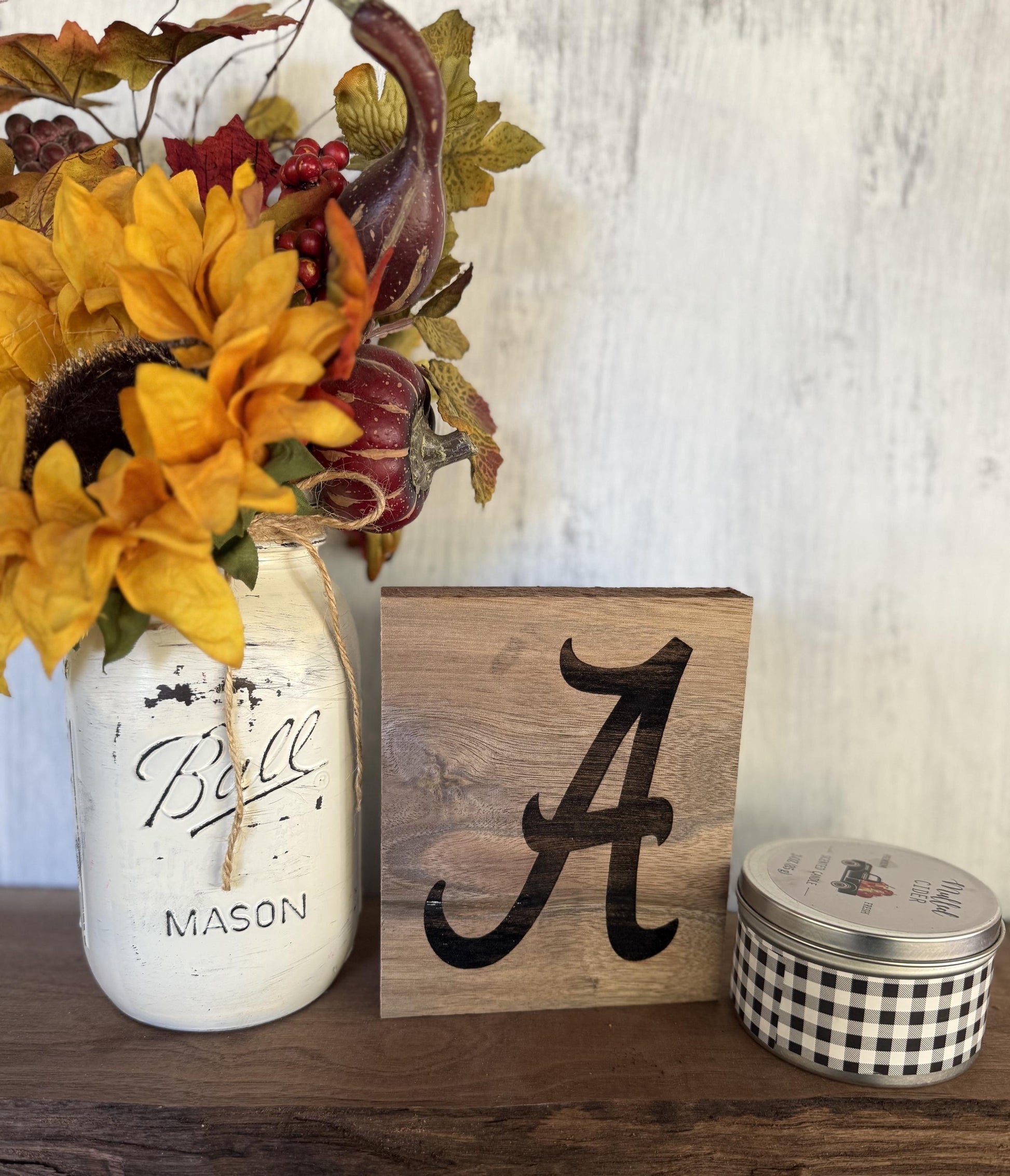 Decorative setup with a mason jar, wooden block with University of Alabama logo and small container on a wooden surface.