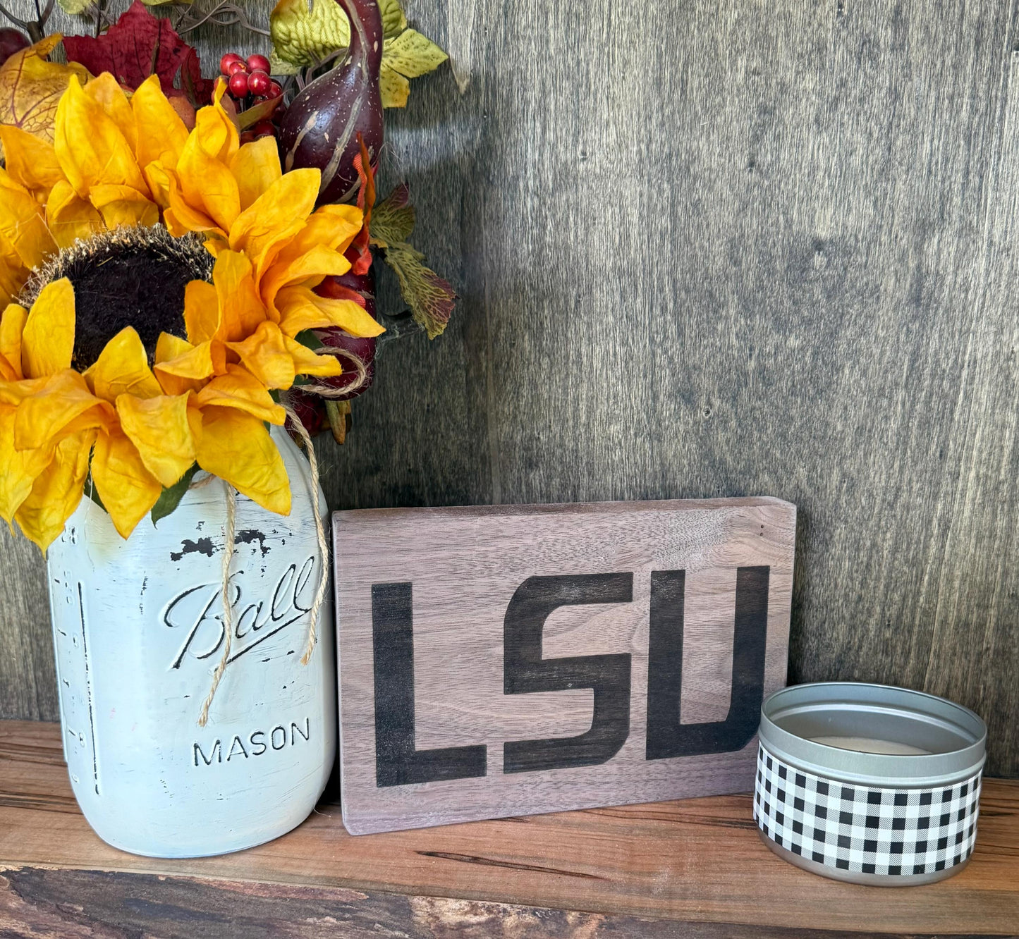 Decorative setup with sunflowers in a mason jar, 'LSU' sign, and small candle on a wooden surface.